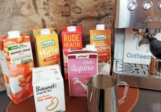 Primer plano de varios tetrabriks de bebidas veganas junto a una jarra de leche y una cafetera espresso