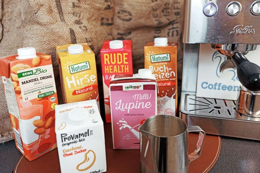 Primer plano de varios tetrabriks de bebidas veganas junto a una jarra de leche y una cafetera espresso