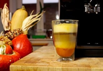 Vaso de pumpkin spice latte junto a unas calabazas y a una cafetera superautomática