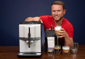 Arne posa sonriente en la prueba de la cafetera superautomática Siemens EQ 300, con un paquete del café de Coffeeness y con algunas bebidas recién preparadas