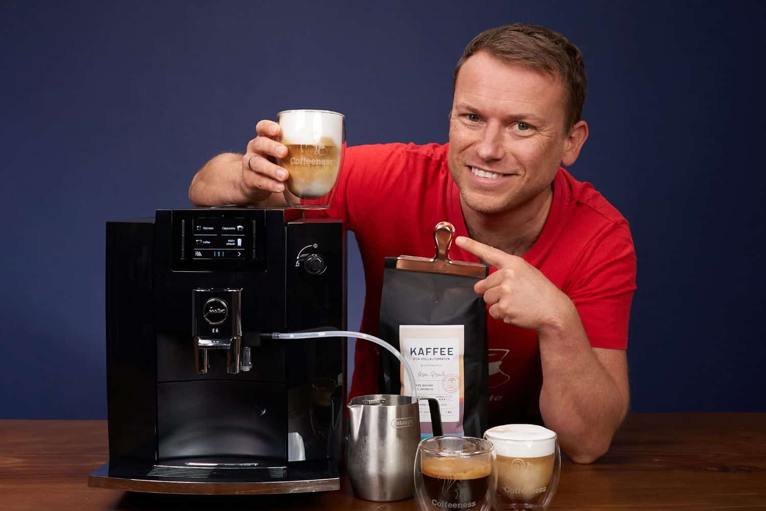 Arne posa sonriente con la cafetera superautomática Jura E6, con un latte macchiato en la mano, otras bebidas recién hechas y un paquete de café de Coffeeness