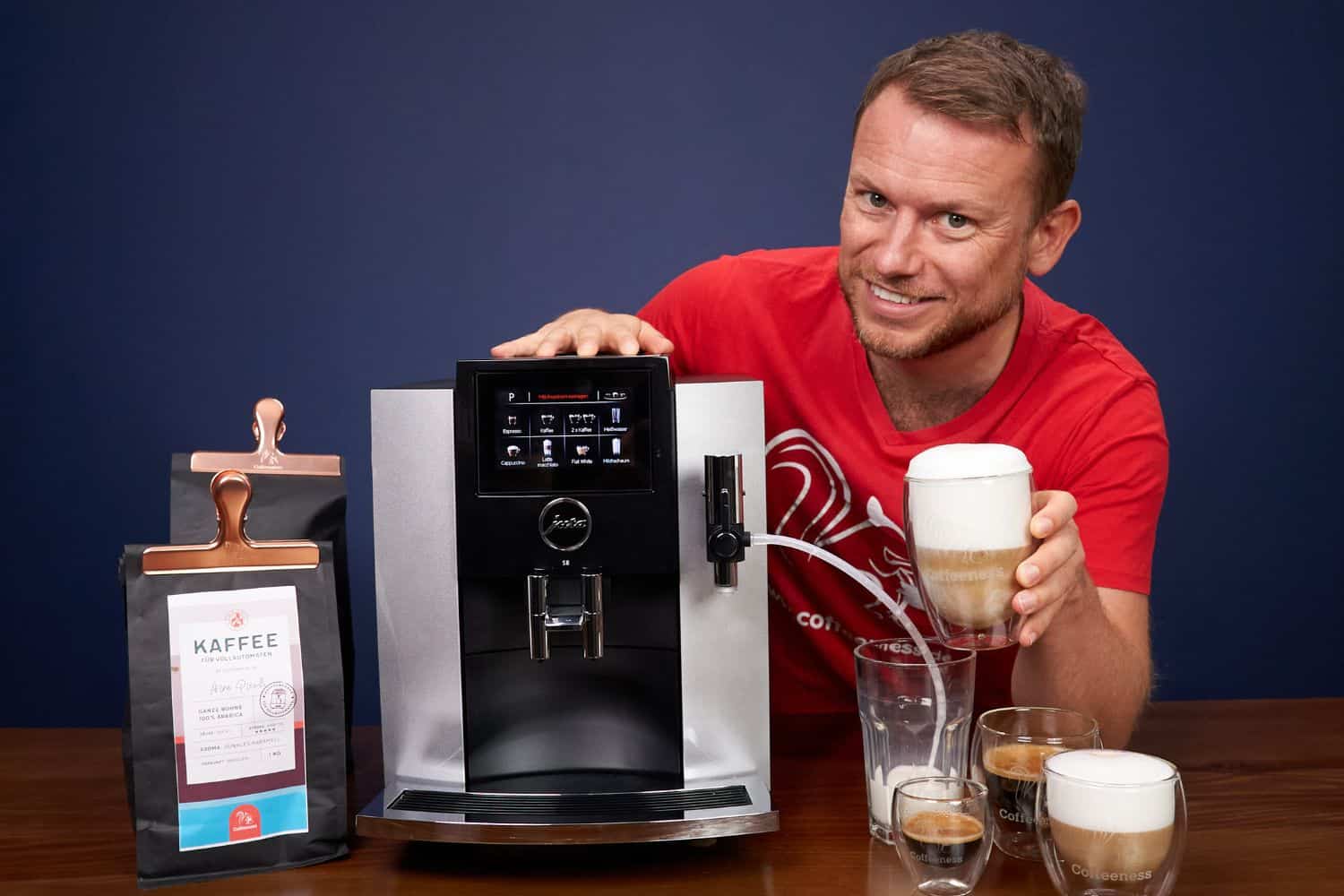 Arne posa sonriente con la cafetera superautomática Jura S8, un latte macchiato en la mano y otras bebidas preparadas con el café de Coffeeness