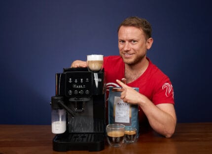 Arne posa sonriente con la cafetera superautomática DeLonghi Magnifica Start (ECAM222.60.BG) y señala con el dedo un capuchino preparado con el café de Coffeeness