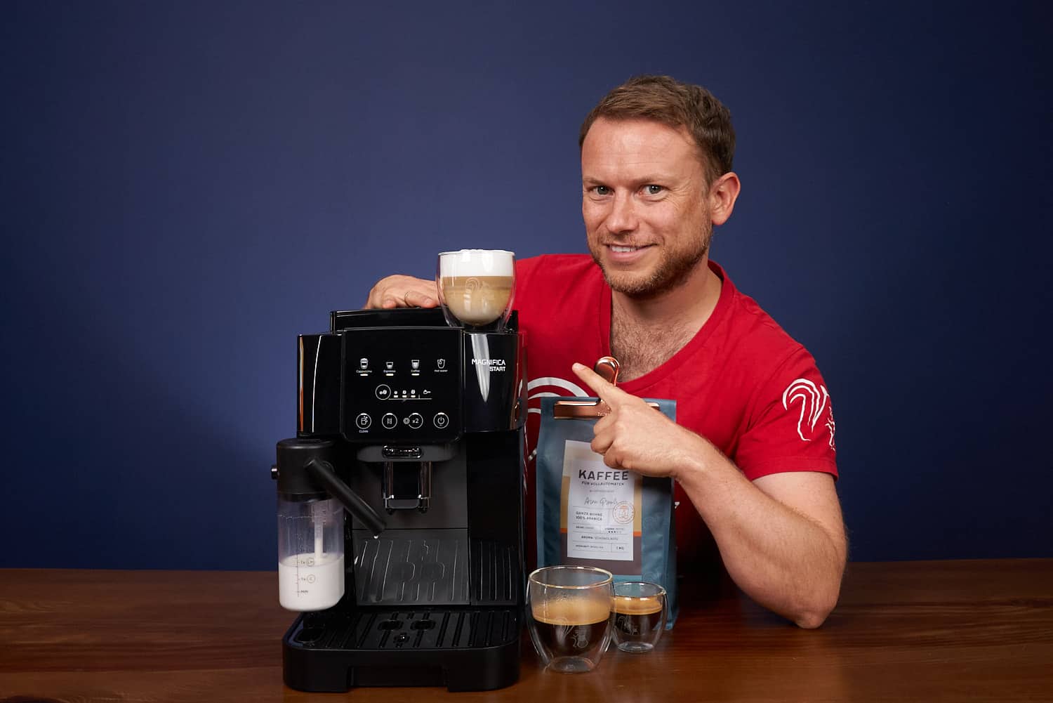 Arne posa sonriente con la cafetera superautomática DeLonghi Magnifica Start (ECAM222.60.BG) y señala con el dedo un capuchino preparado con el café de Coffeeness
